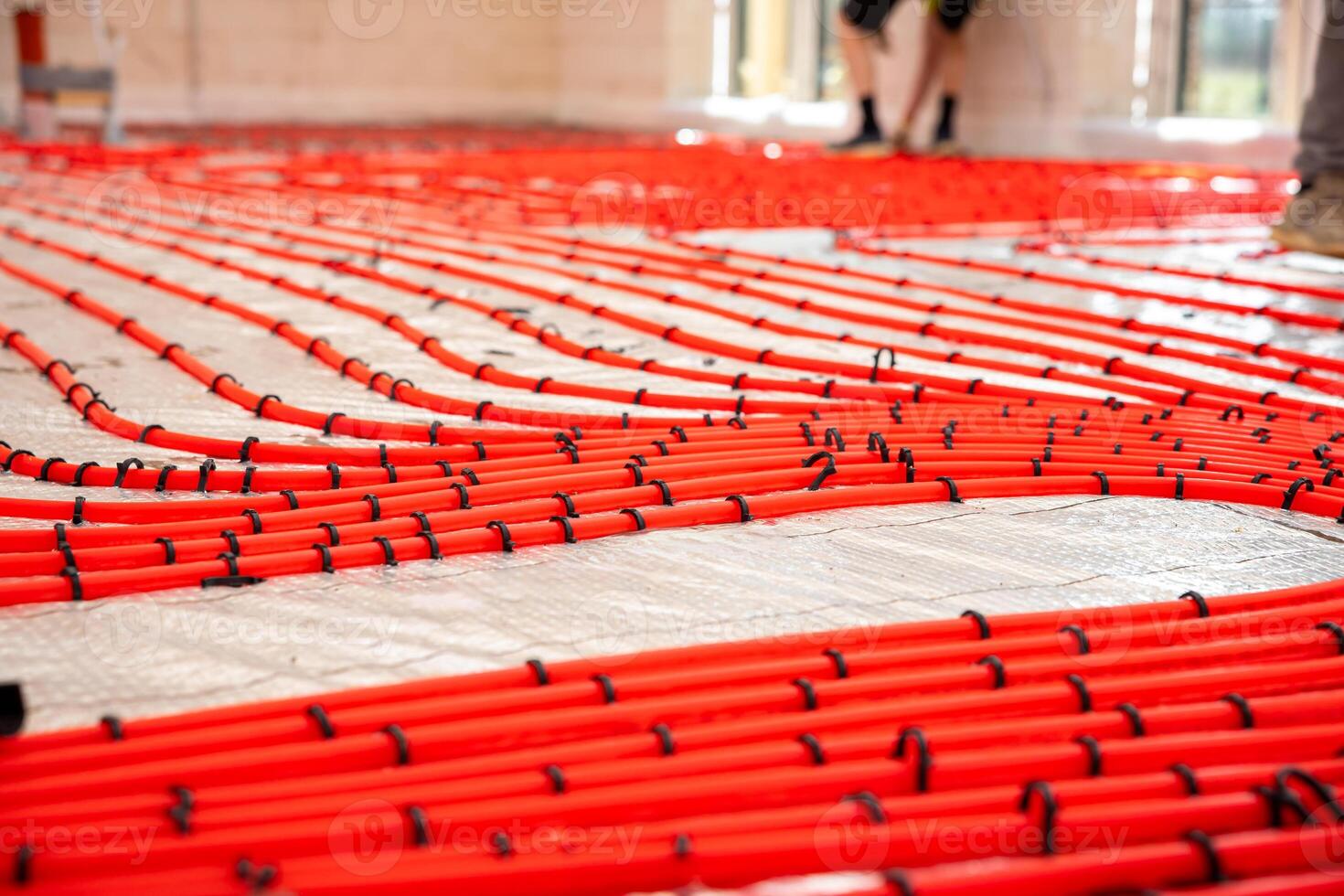 Bright red heating pipes arranged in a grid on reflective insulation, secured with black fasteners. Light walls, large windows, and workers' legs visible. photo