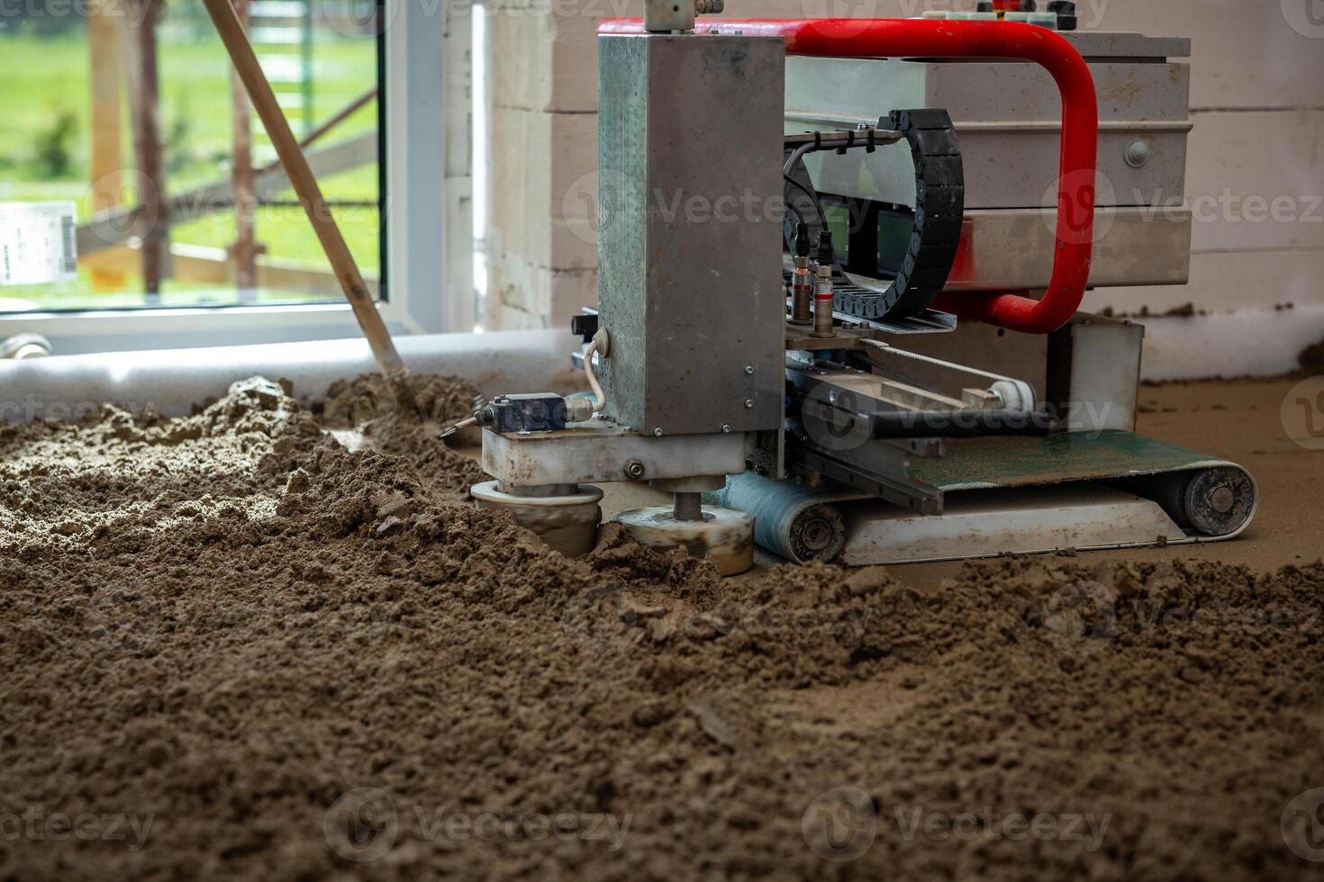 A concrete leveling machine with a red handle operates on wet material indoors. Natural light enters through a large window, revealing blurred green surroundings. photo