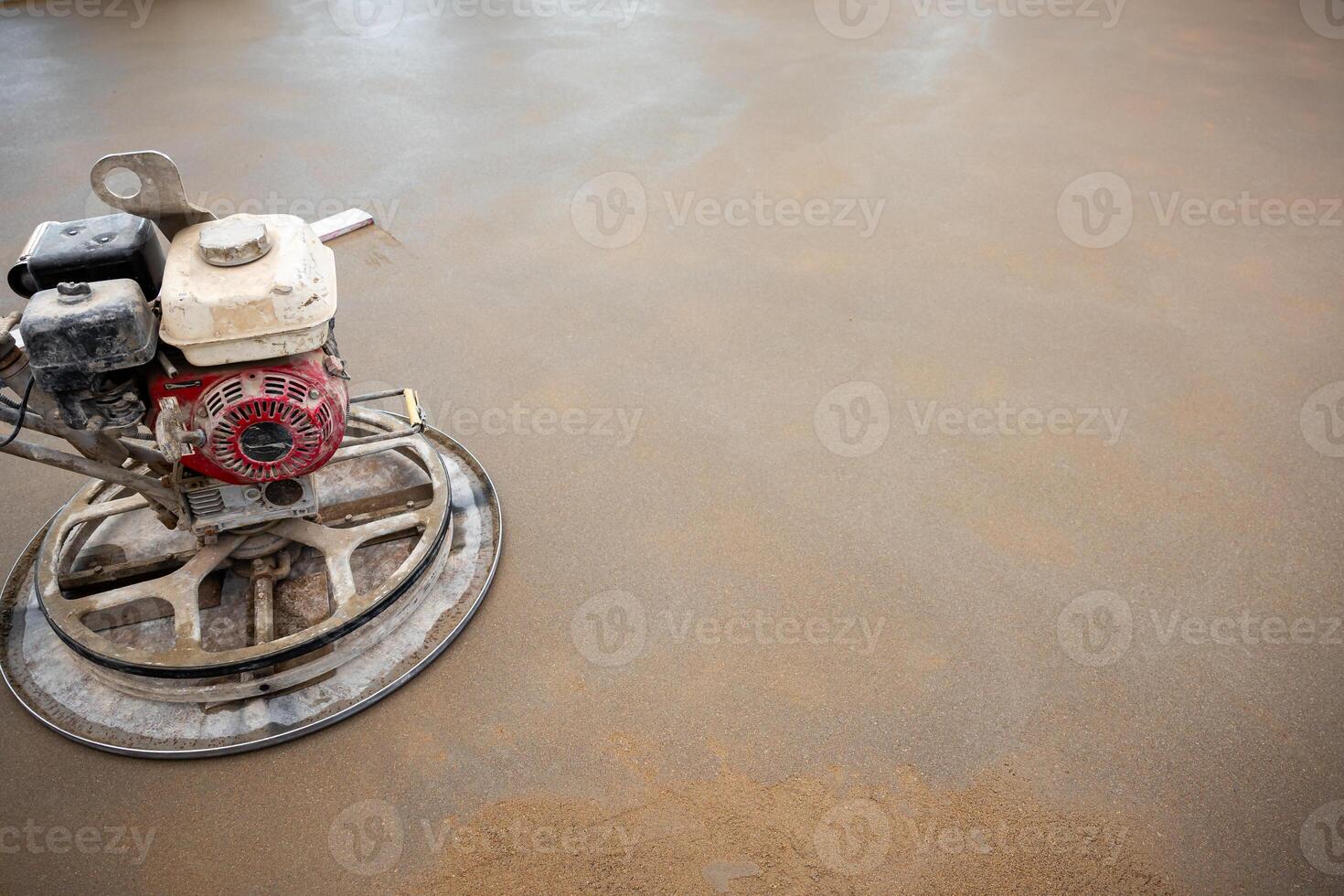 A power trowel machine with a red and white motor smooths a wet concrete surface. The circular blade contacts the floor, showing drying patches. photo