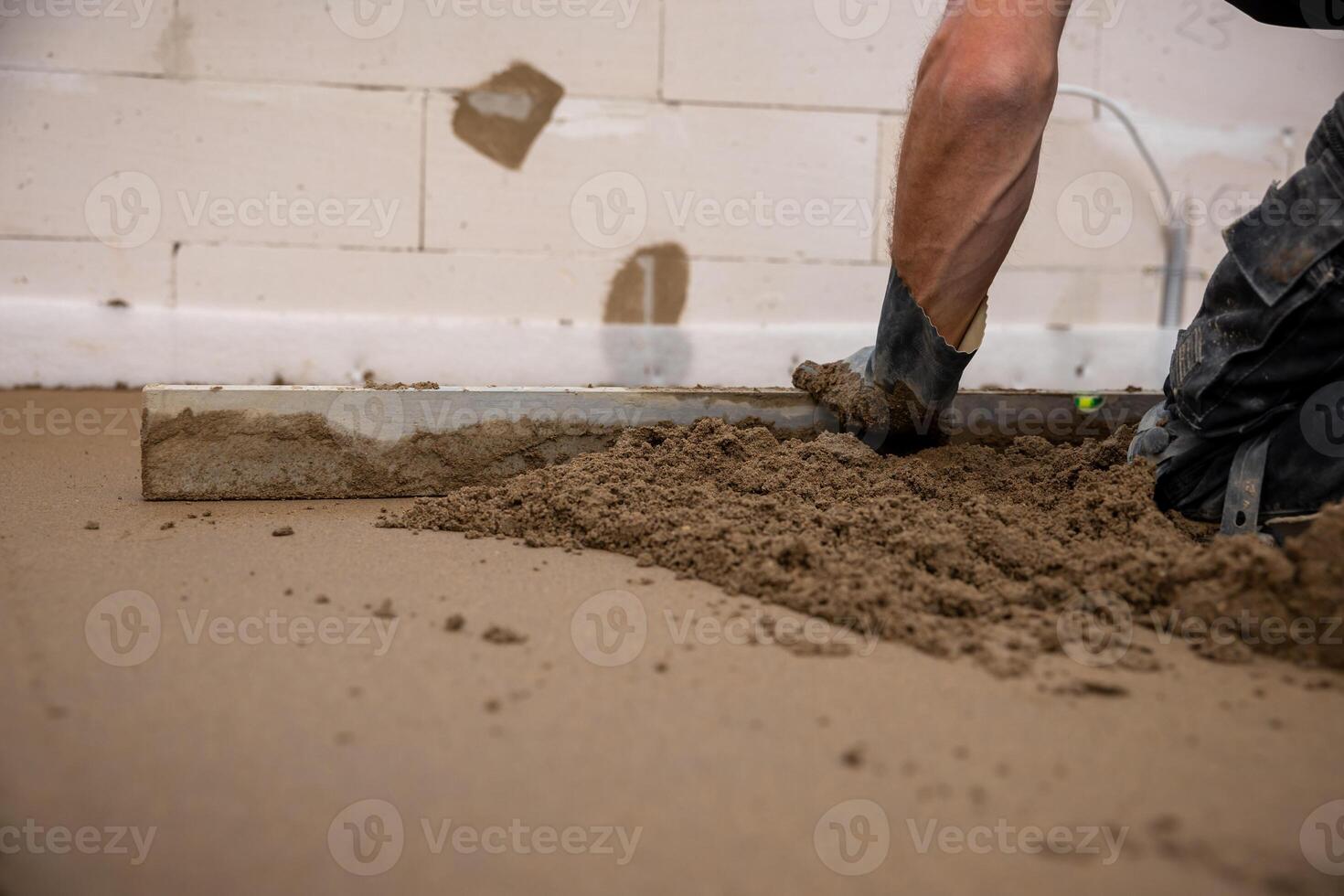 A worker uses a leveling tool to smooth Estrich concrete over underfloor heating pipes. Partially finished walls and exposed material are visible indoors. photo