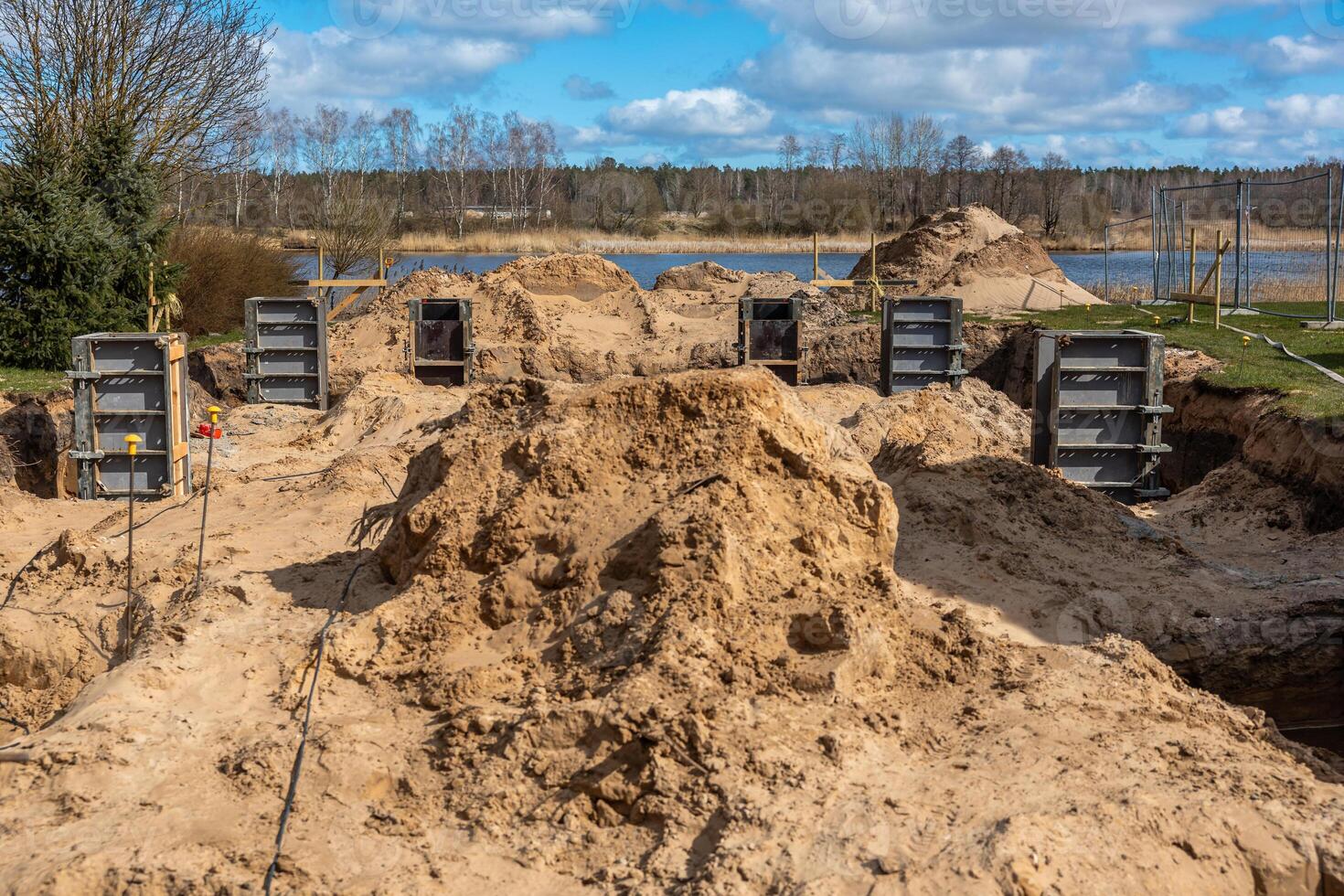 Active construction site featuring sandy excavations, metal framework, and fenced off area. A body of water with sparse trees and reeds is visible. photo
