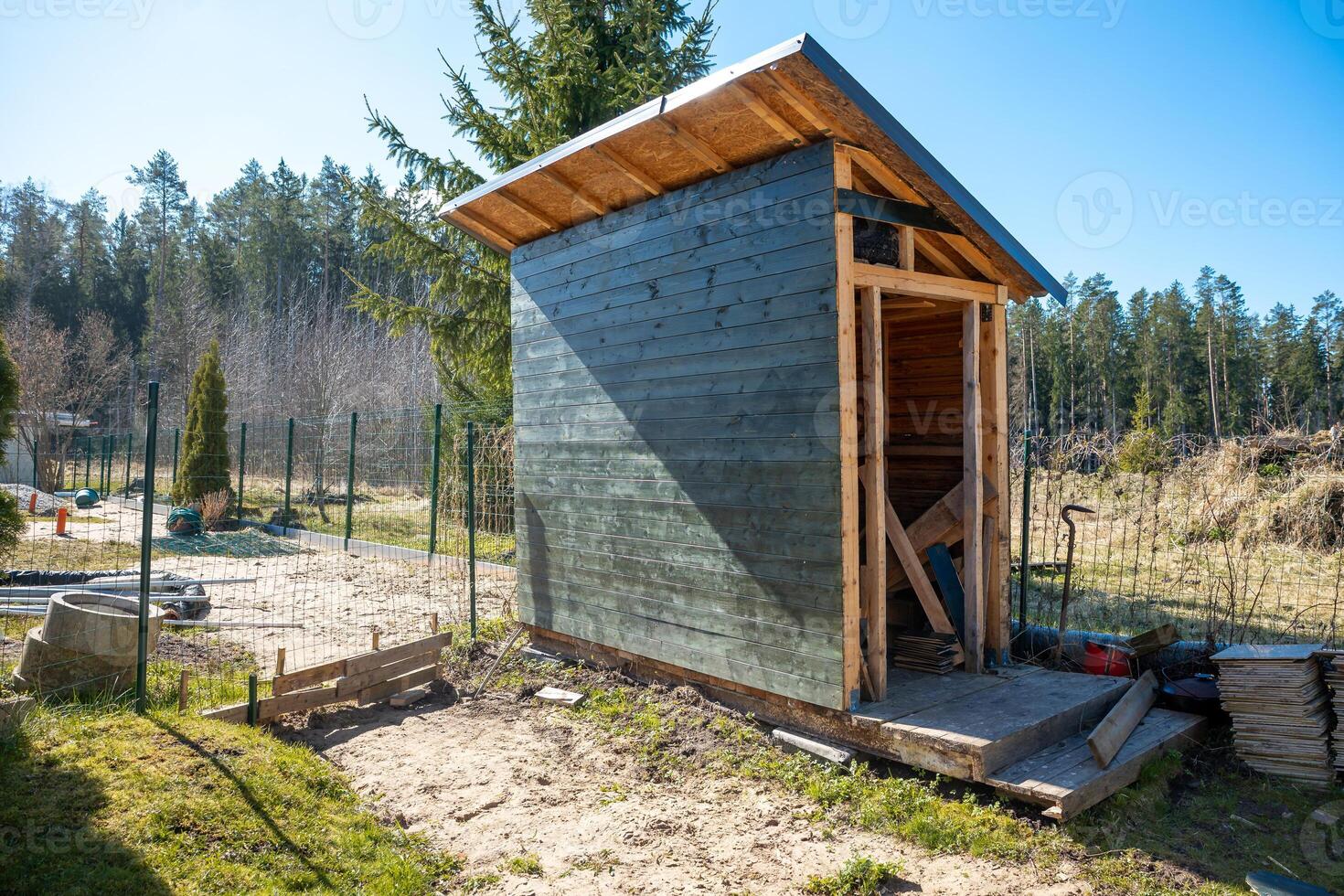 A small wooden shed with a slanted roof, partially painted green, surrounded by a wire fence. Tools and materials lie nearby under bright sunlight. photo