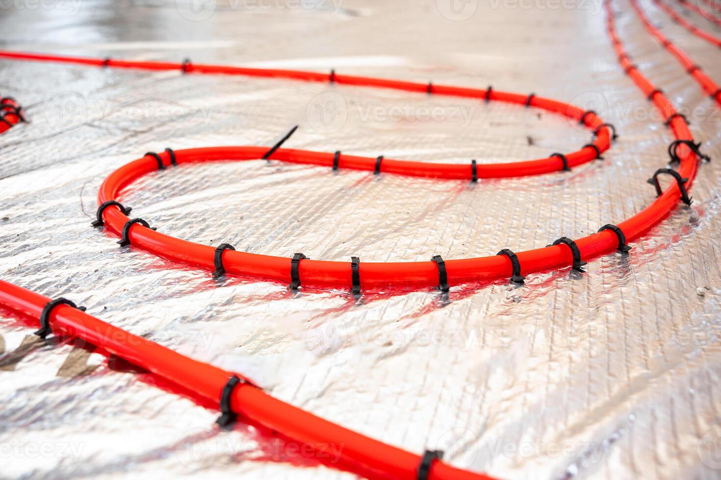 Bright red heating pipes are coiled and secured with black fasteners on a reflective foil insulation layer, highlighting the installation process. photo