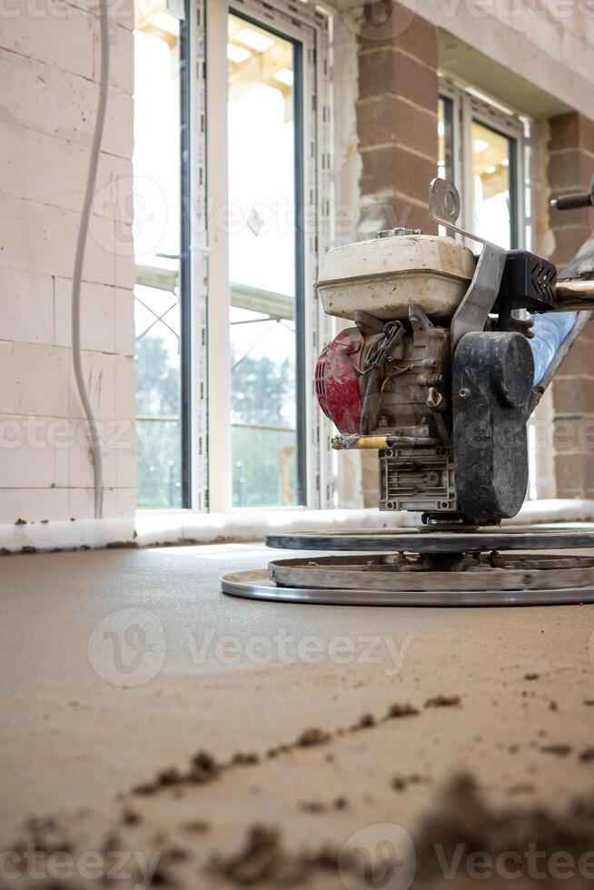 Interior of a construction site showing a power trowel smoothing concrete flooring over underfloor heating pipes, with brick walls and large windows. photo