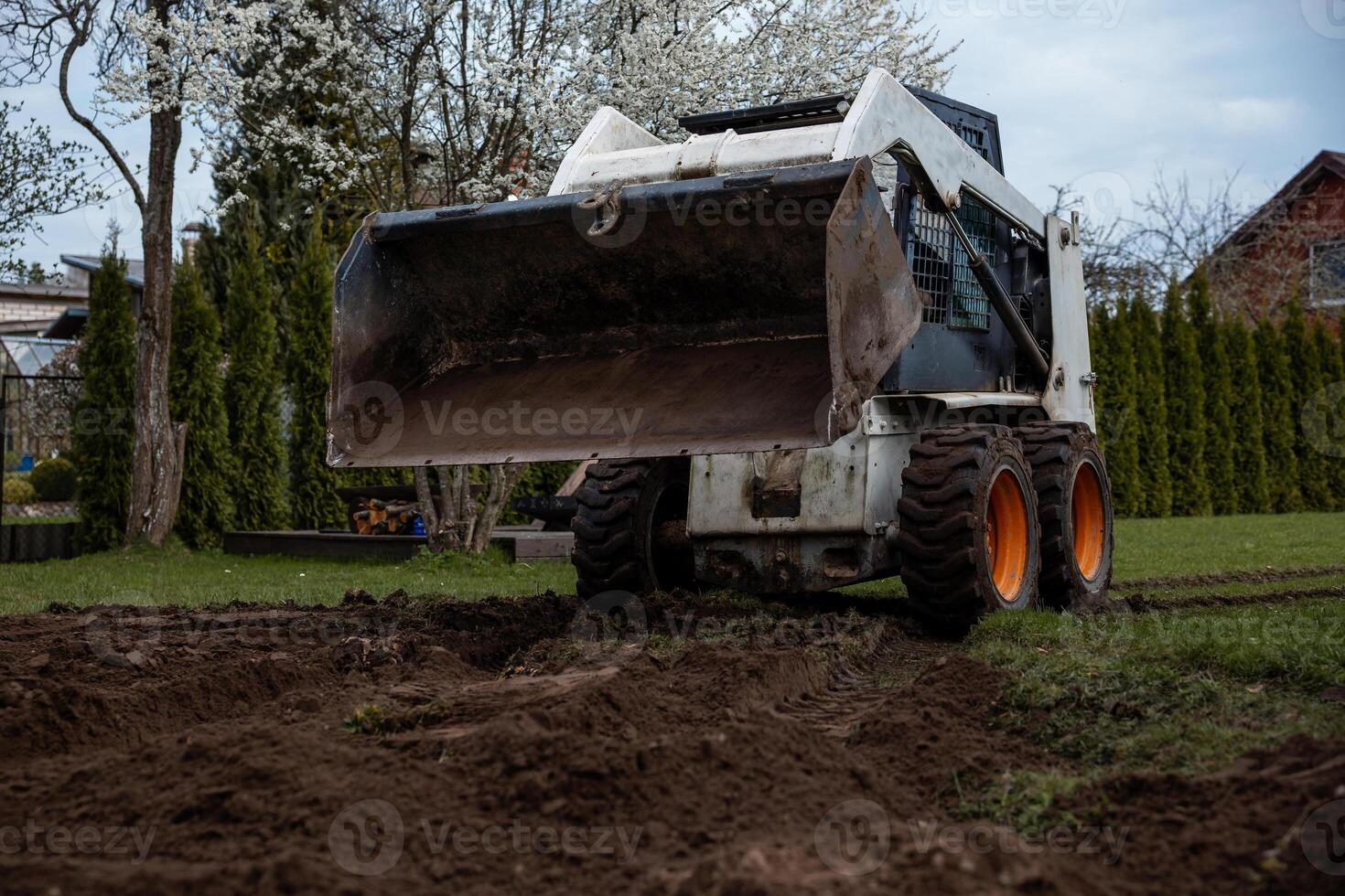 A skid steer loader with orange wheels rests on freshly dug soil near trees with white blossoms, trimmed hedges, and a house with a sloped roof. photo