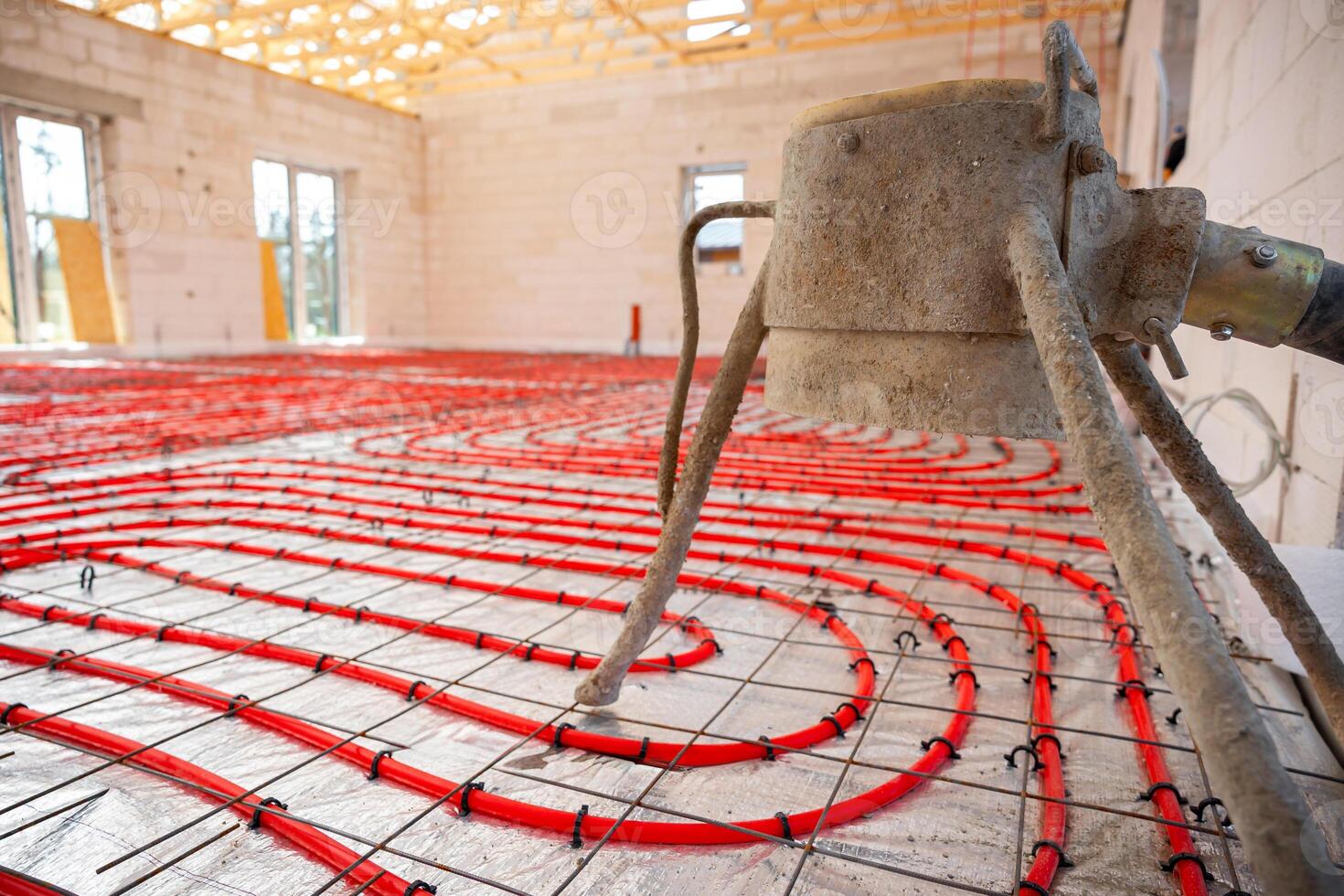 Interior of a construction site showing coiled underfloor heating pipes on a metal grid, a concrete mixer, light brick walls, and exposed wooden beams. photo