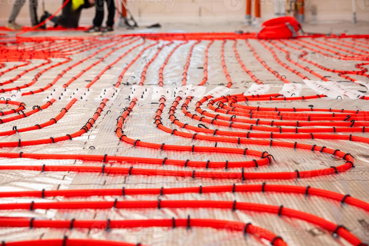 Bright red underfloor heating pipes arranged in coiled patterns on a reflective surface, with workers in high visibility clothing in the background. photo