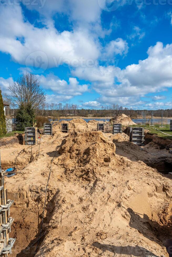 A construction site featuring sand piles, metal framework, and concrete forms. Open land with sparse trees and a distant body of water are visible. photo