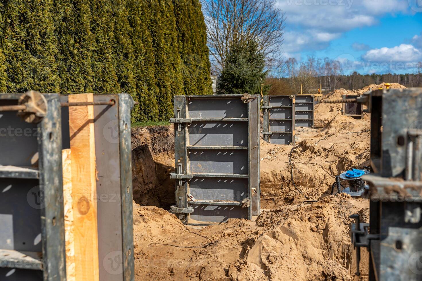 Metal framework panels stand on sandy soil at a construction site. A tall hedge borders one side, with leafless trees and sparse vegetation visible. photo