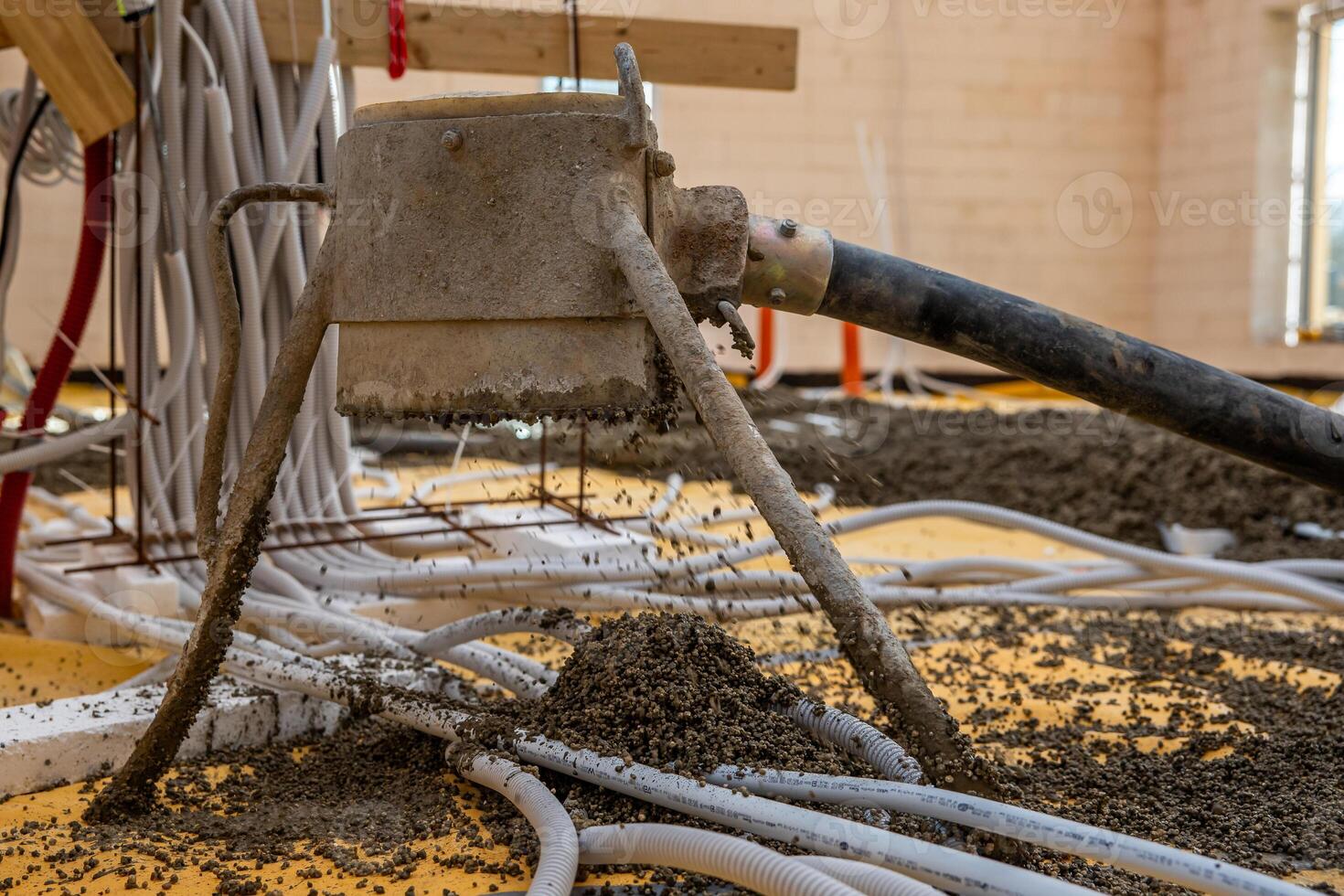 Concrete pouring machine dispenses material on a yellow base layer with rebar. Scattered cables, pipes, and unfinished walls indicate early construction work. photo