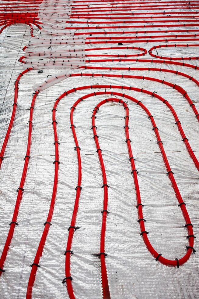 Red underfloor heating pipes arranged in loops, secured with black fasteners on a reflective insulation layer, part of a construction process. photo