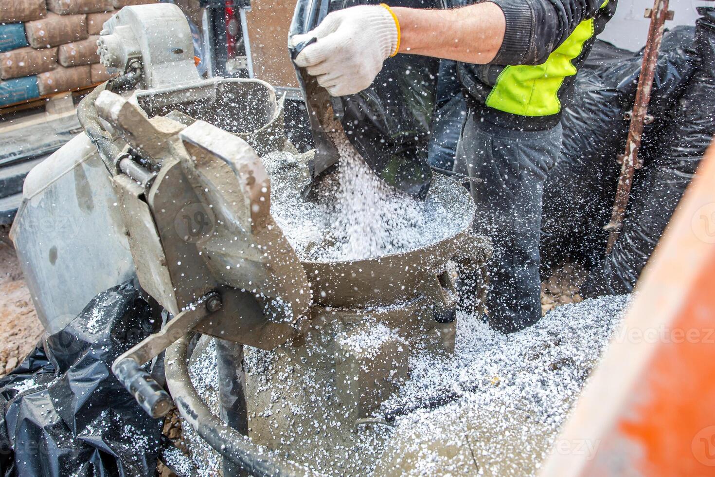 A worker in high visibility gear pours white granules into a concrete mixer surrounded by black coverings and construction materials at an active site. photo
