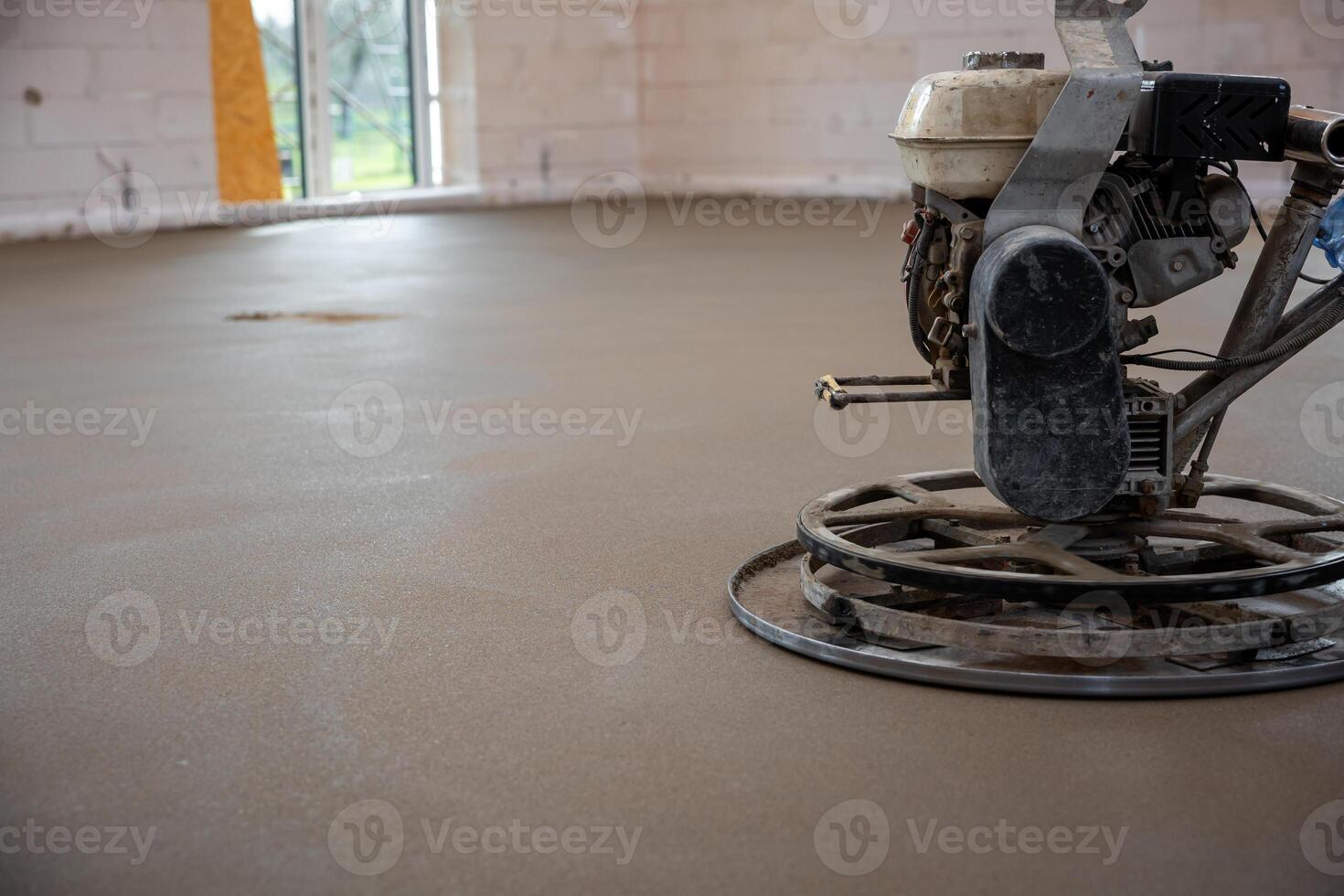 Construction site interior with a power trowel smoothing a concrete floor. Unfinished walls with white bricks and an open window provide natural light. photo