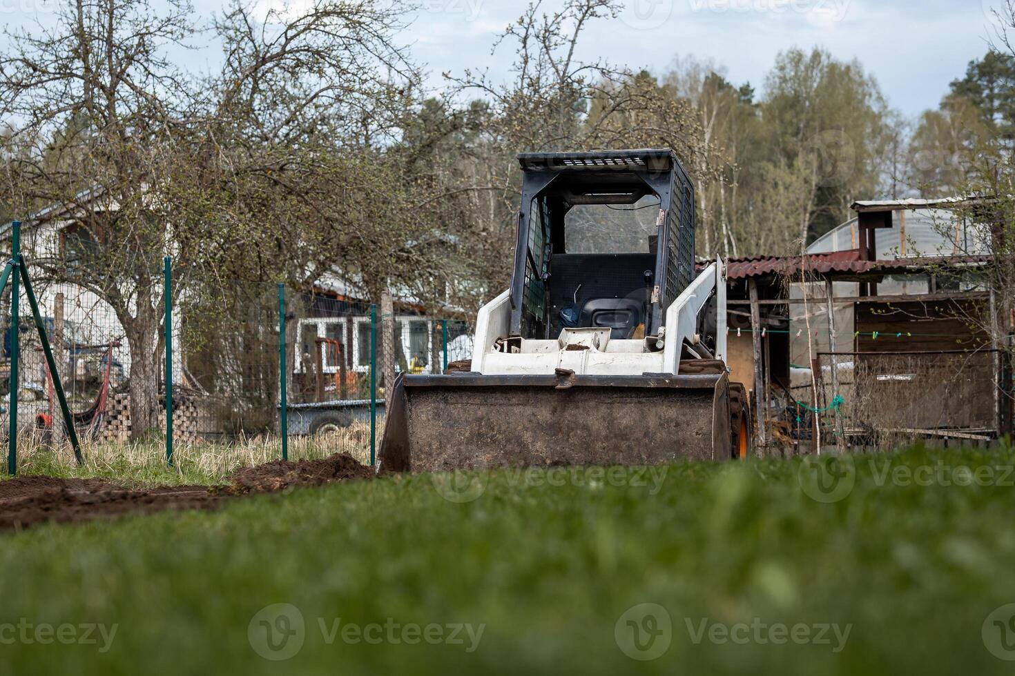 A white skid steer loader with its bucket lowered sits on grass, surrounded by sparse trees, a fenced area, a shed, and a house in a rural setting. photo