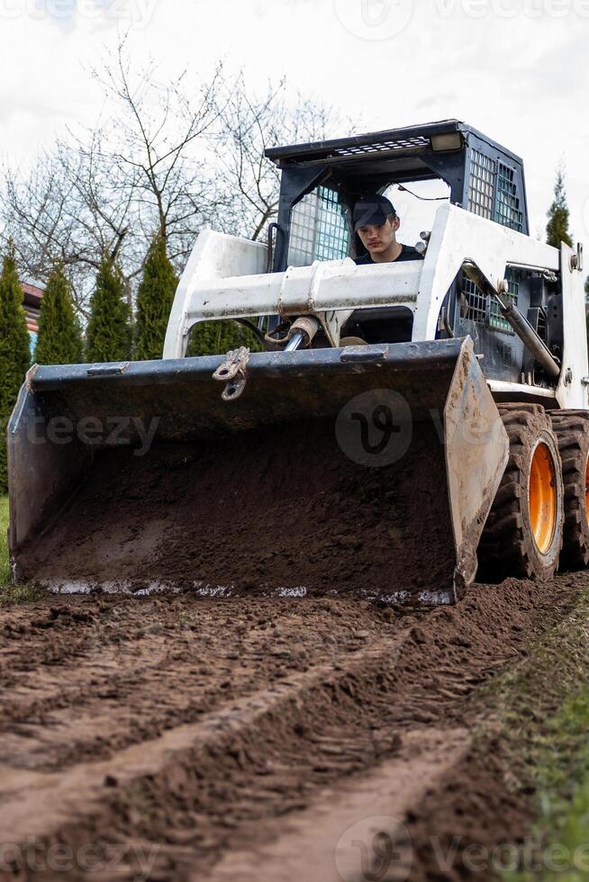 A skid steer loader with orange wheels moves soil in an outdoor area. Tall conical trees and bare branches are visible in the distance. photo