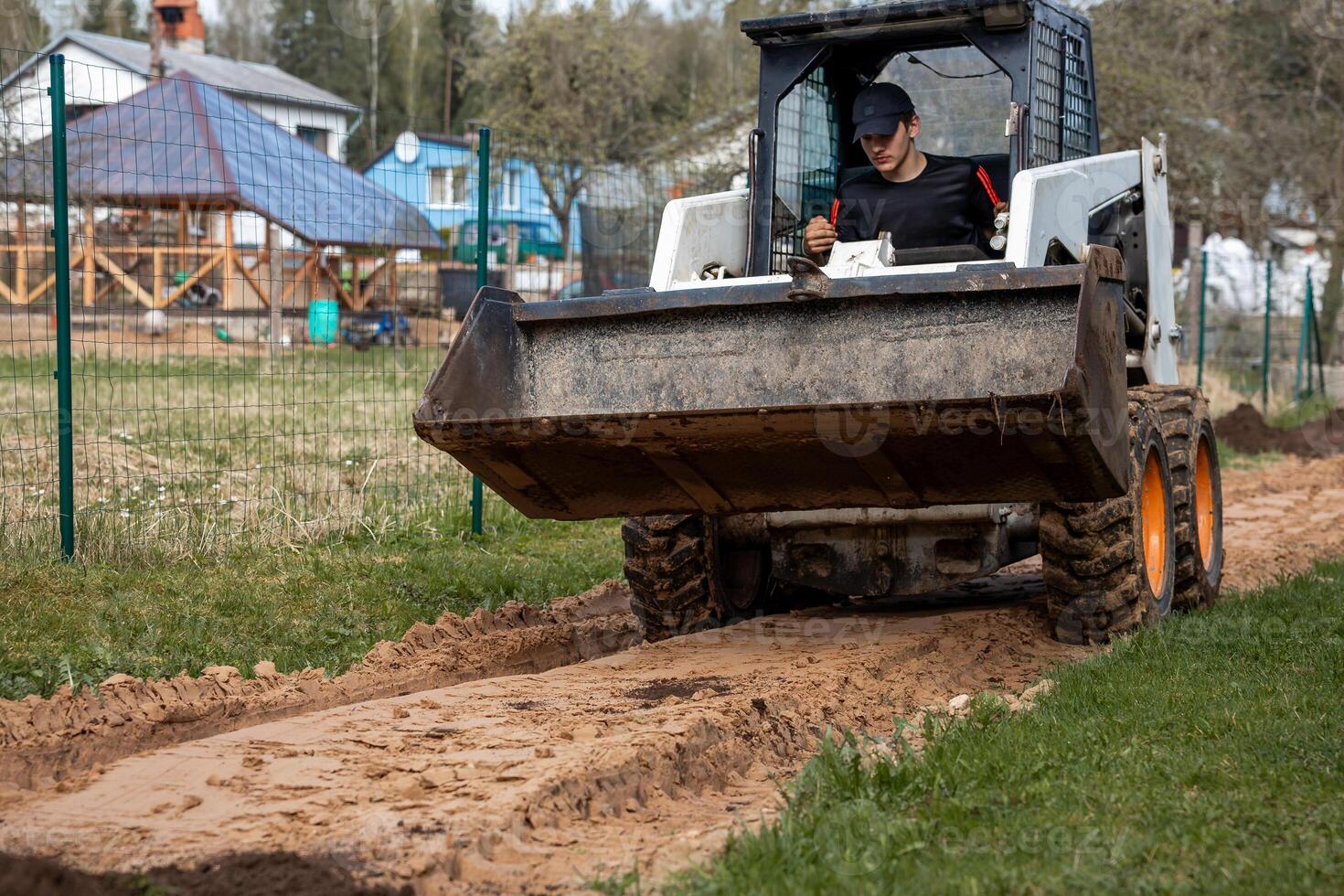 A man uses a skid steer loader with a front bucket to level a dirt path. Green wire fence and residential houses with pitched roofs are visible nearby. photo