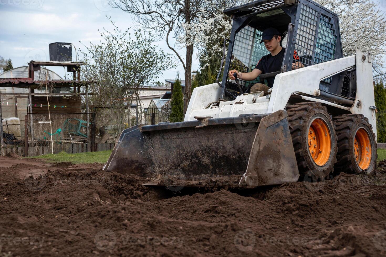 A man uses a skid steer loader with orange wheels to move soil. The area includes trees, some in bloom, a shed, fencing, and natural surroundings. photo