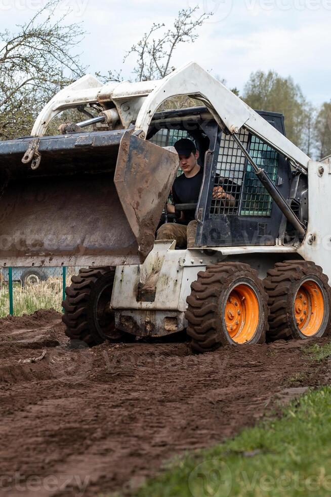 A skid steer loader with orange wheels carries soil in its raised bucket. A person operates the machine in a grassy field with sparse trees nearby. photo
