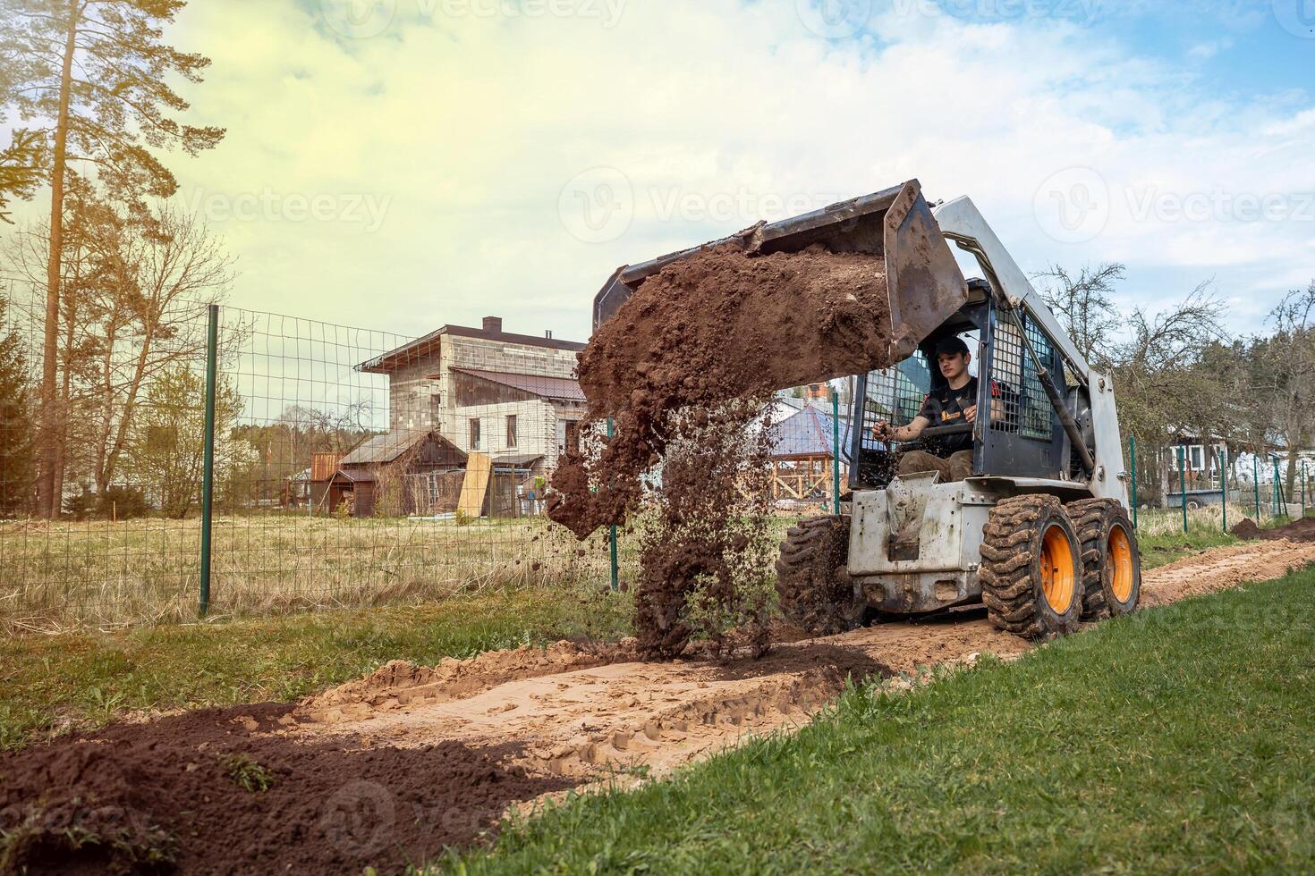 A skid steer loader with orange wheels dumps soil on a pathway near a wooden house with a slanted roof, surrounded by fields and sparse trees. photo