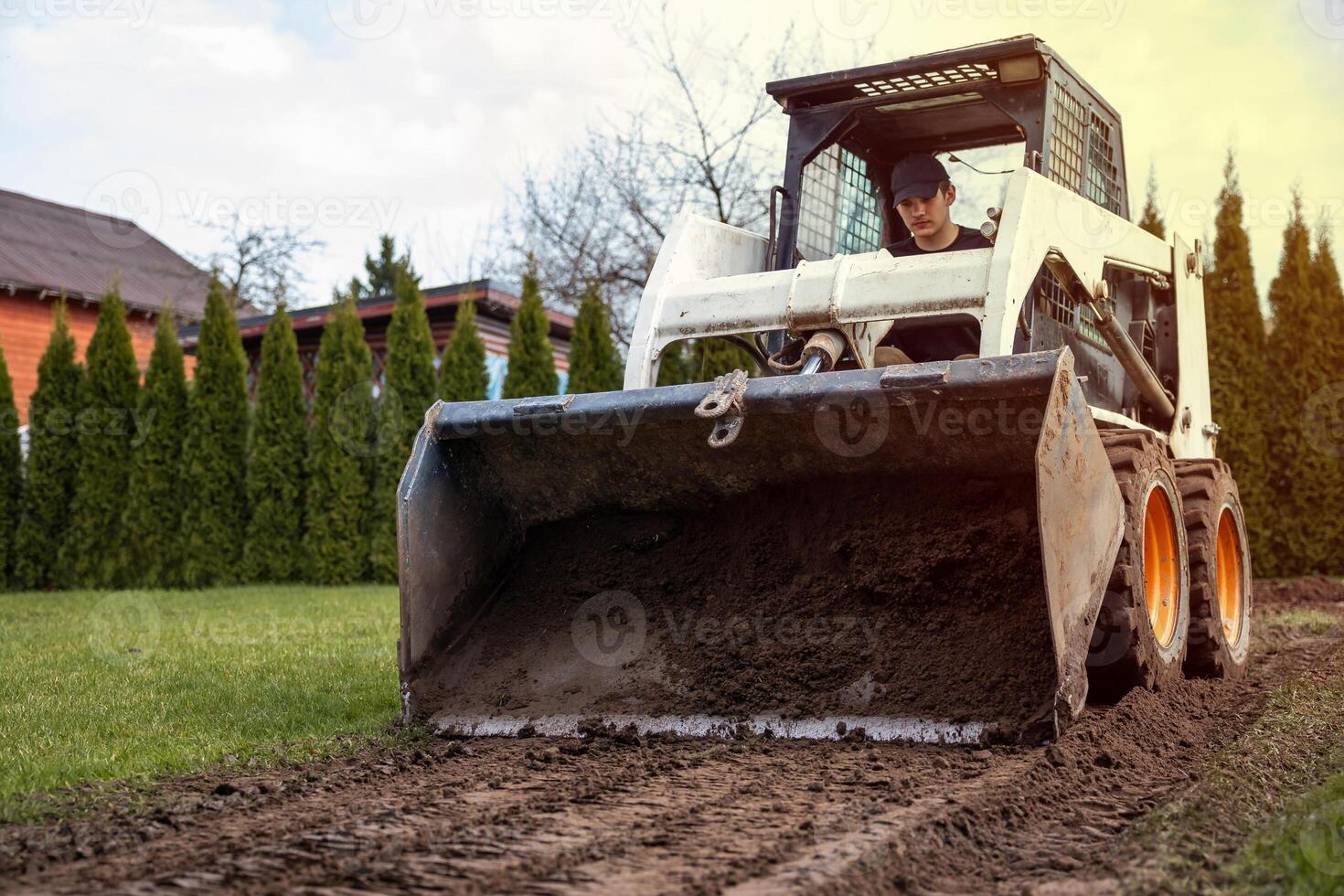 A white skid steer loader with orange wheels levels soil on a dirt path. A person operates it near tall evergreen trees and a slanted roof structure. photo