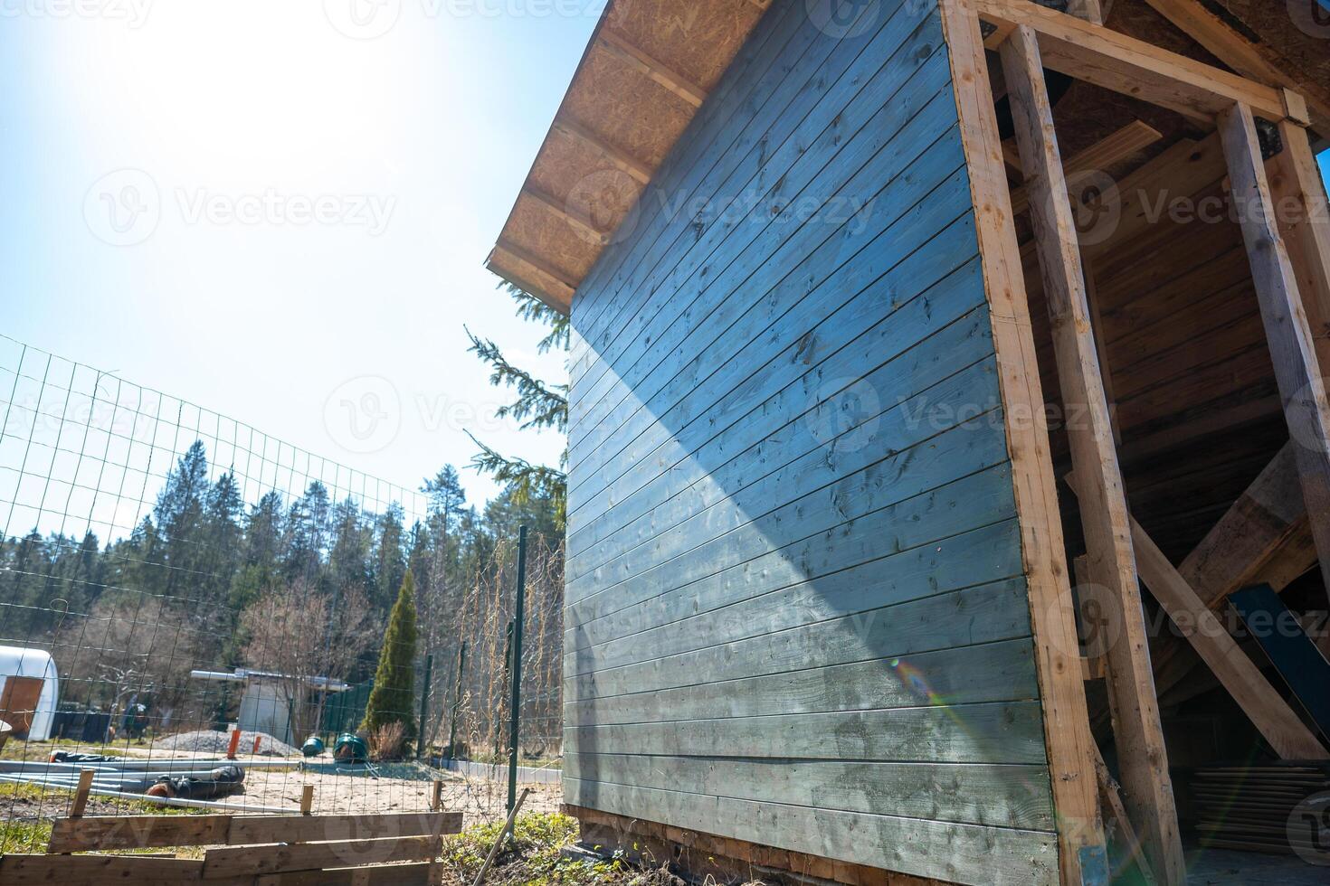 A blue wooden structure with a slanted roof stands near a wire fence. Scattered objects and a small structure are visible, with tall trees in the background. photo