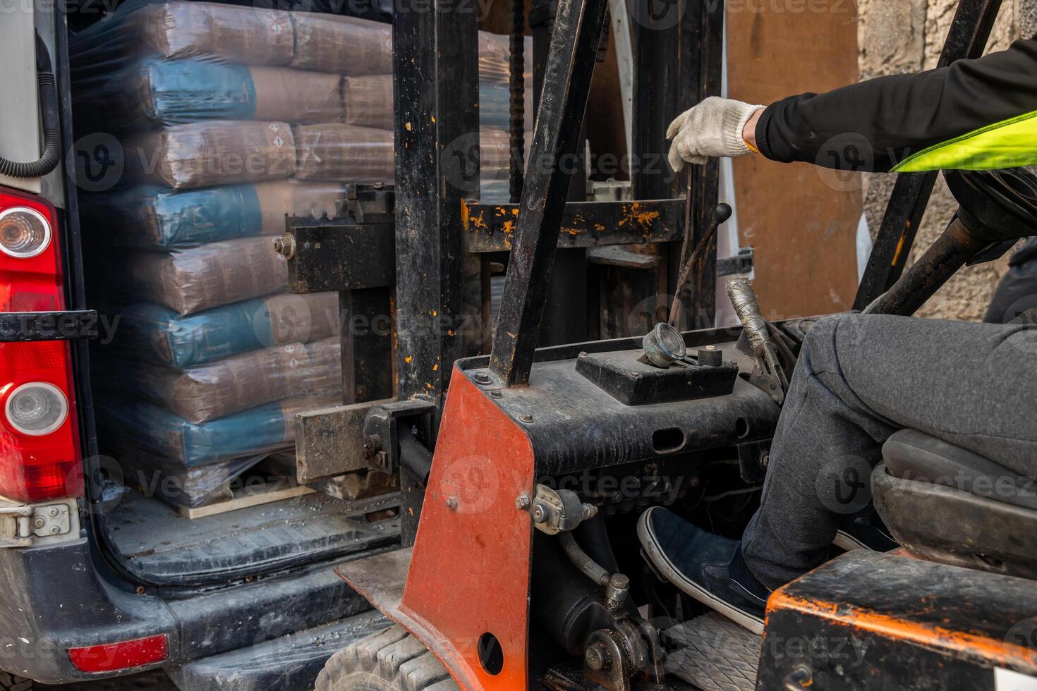 A worker in high visibility gear operates a forklift to load or unload plastic wrapped goods from a vehicle. A stone wall is partially visible nearby. photo