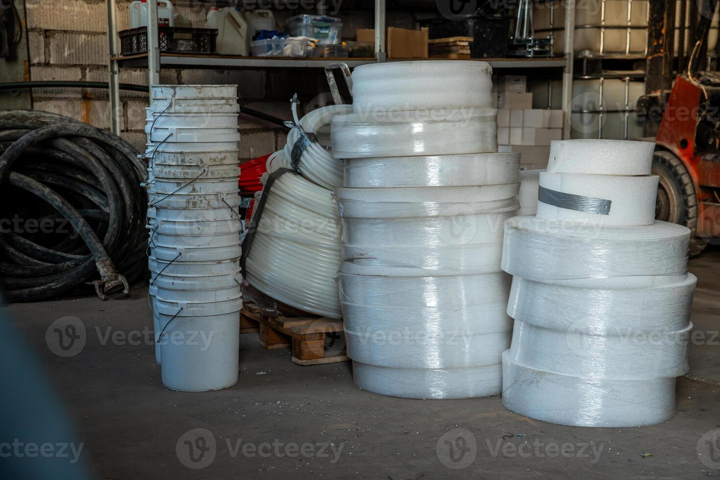 Concrete floor workspace featuring stacked white buckets, foam rolls, coiled hoses, tools, shelves with supplies, and a partially visible forklift. photo