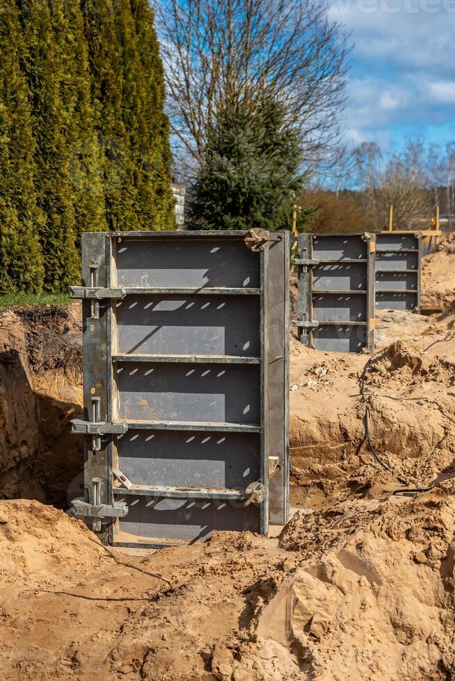 Metal framework panels rest in sandy soil at a construction site, surrounded by evergreen trees and a leafless deciduous tree, with visible trenches. photo