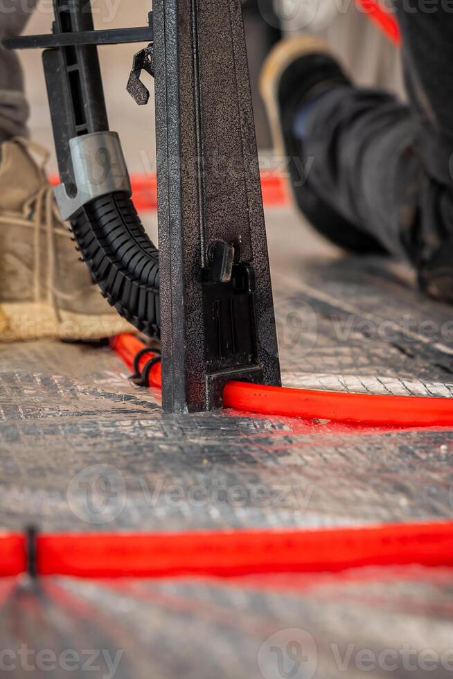Red underfloor heating pipes are laid on a reflective surface, with a metal tool and a worker's shoe visible, capturing active installation work. photo
