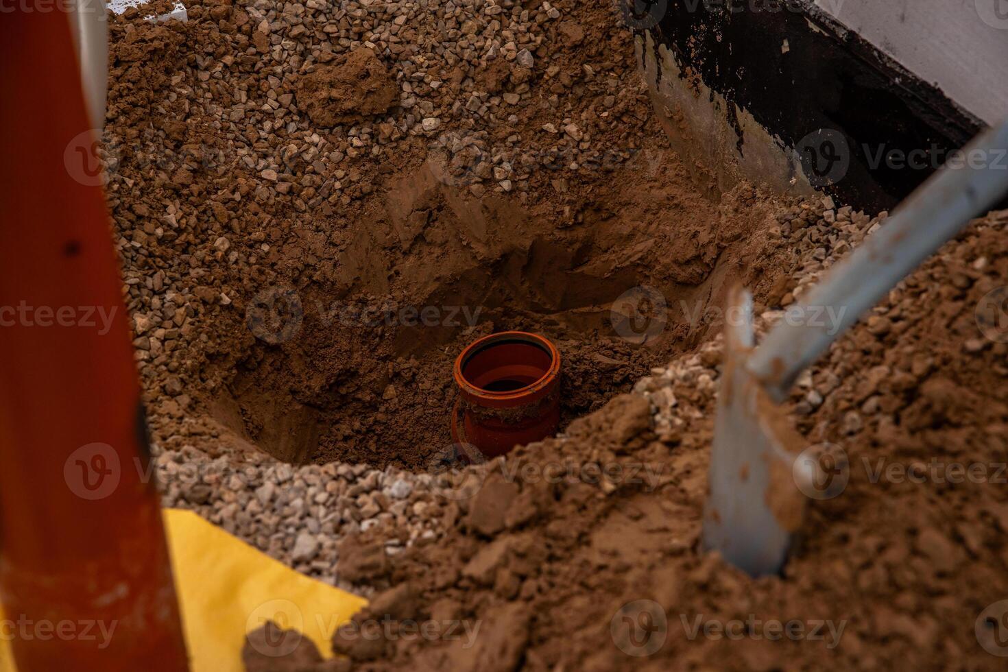 A construction site featuring a partially dug hole with a red brown pipe in the soil, loose dirt, gravel, a shovel, and a concrete wall with black waterproofing. photo