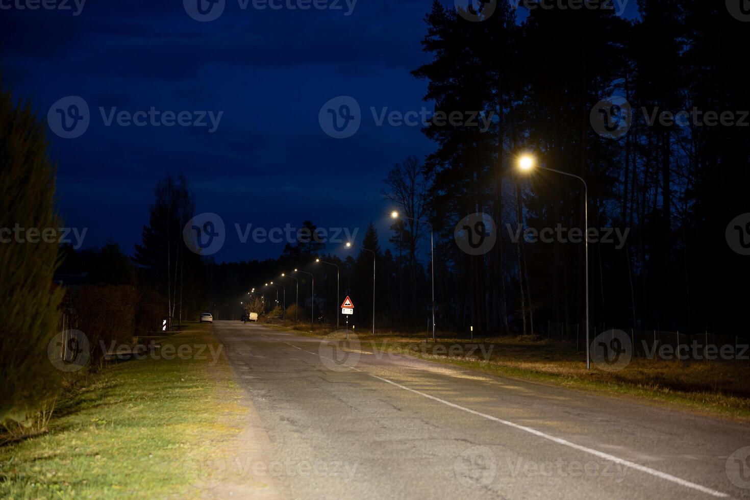 A road extends under a deep blue evening sky, bordered by trees and grass. Streetlights illuminate the pavement, with a triangular traffic sign visible. photo