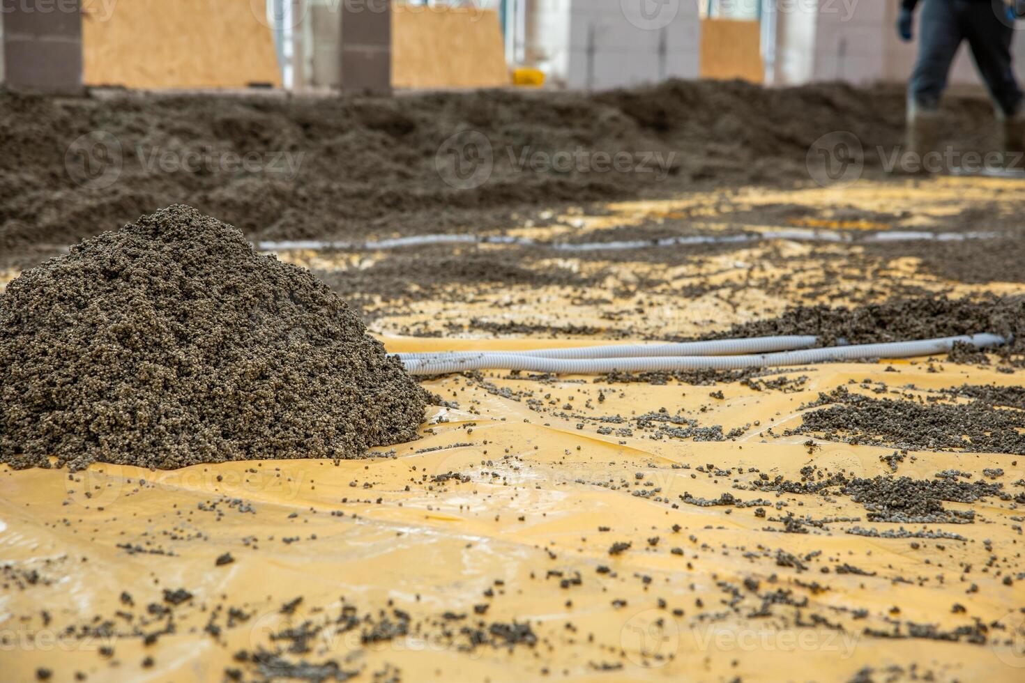 A construction site featuring a gravel pile on a yellow plastic sheet, pipes arranged for foundation work, and partially built concrete walls. photo