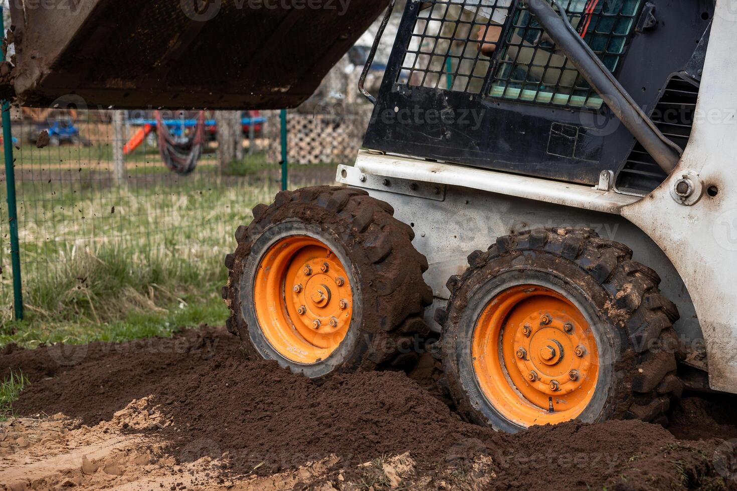 A compact skid steer loader with orange wheels moves soil on uneven ground. Rugged tires and a fenced area with playground equipment are visible. photo