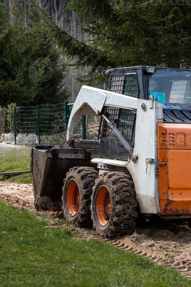 A white and orange skid steer loader with a bucket attachment sits on dirt and grass. A green metal fence and tall evergreen trees are in the background. photo