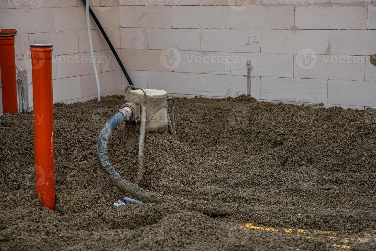 An indoor construction site featuring a floor area under preparation with concrete, two orange vertical pipes, and equipment with a hose in use. photo