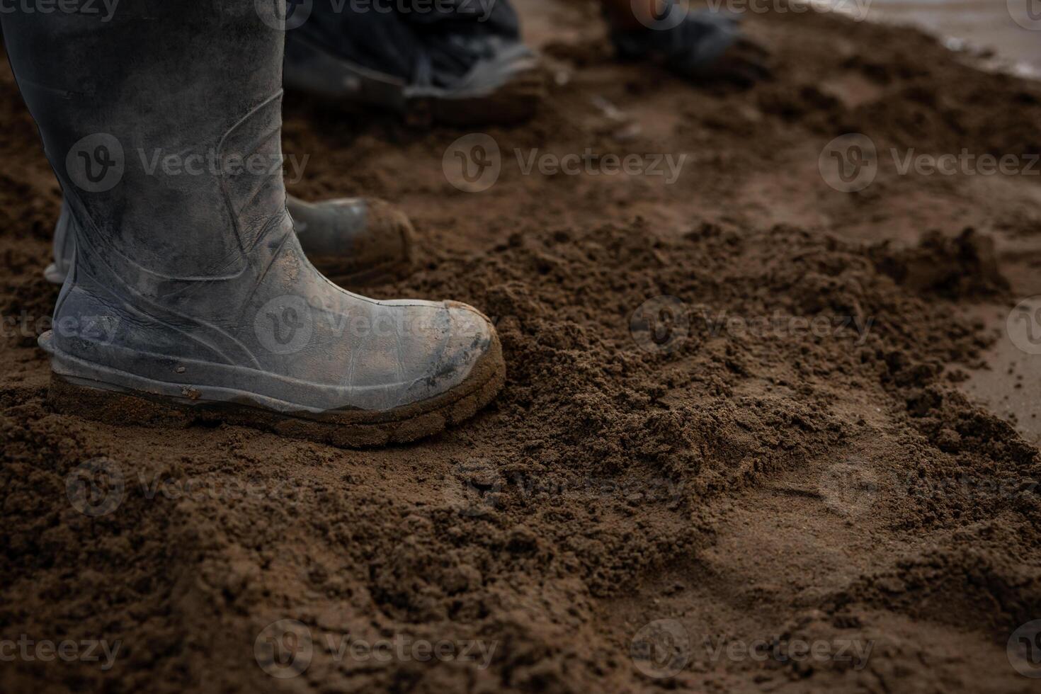 Muddy boots stand on damp, uneven soil with additional footwear in the background. The ground appears freshly disturbed, suggesting outdoor work activity. photo