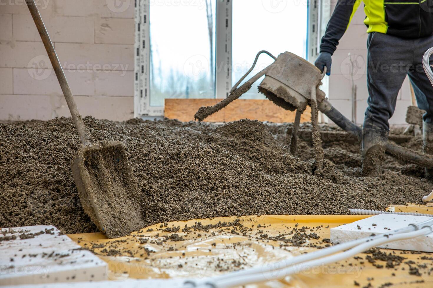 Construction workers pour and level concrete over yellow plastic sheeting and insulation panels, with tools and bare trees visible through window openings. photo