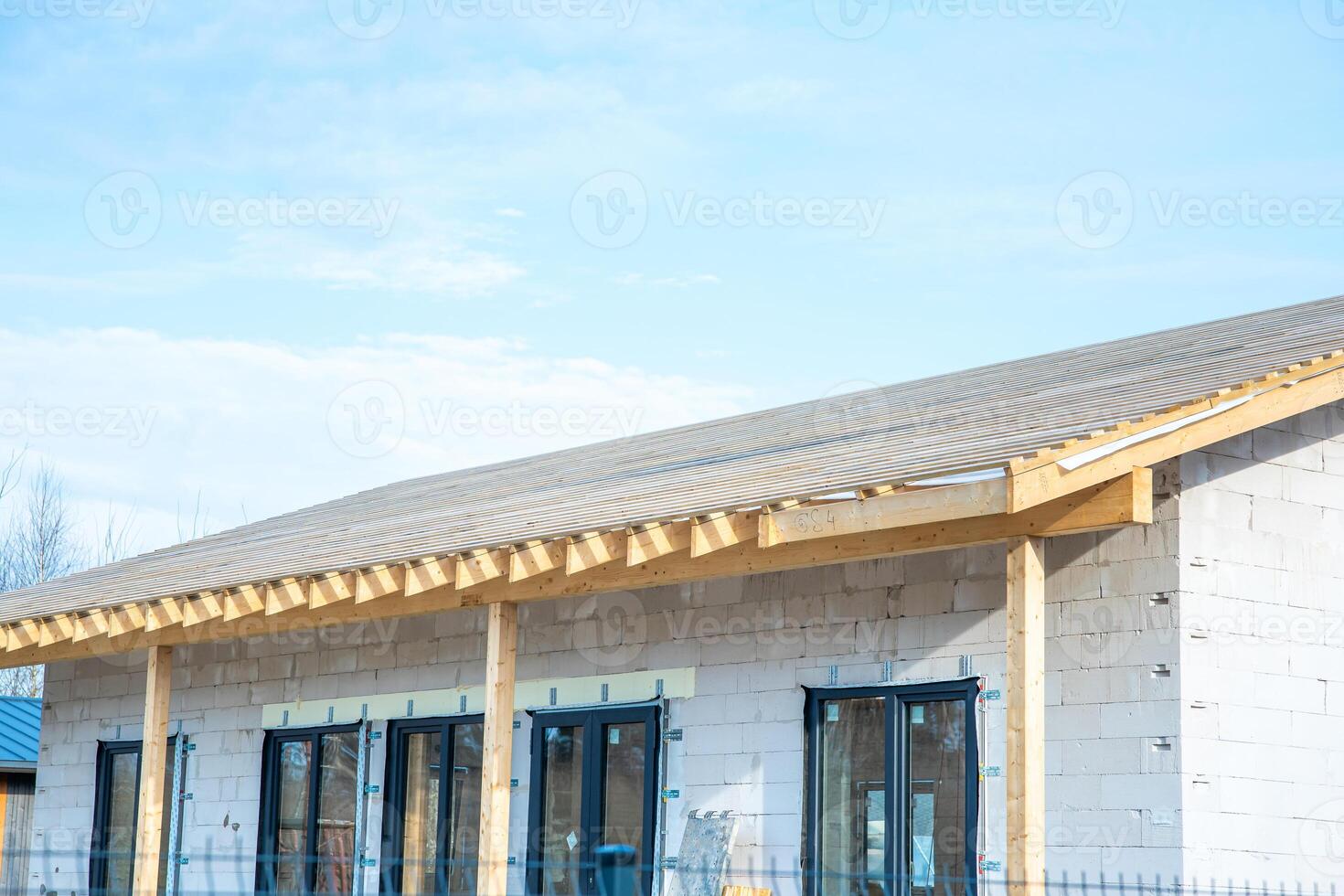 A building under construction with white concrete block walls, dark framed windows, and an unfinished wooden roof frame under a clear blue sky. photo