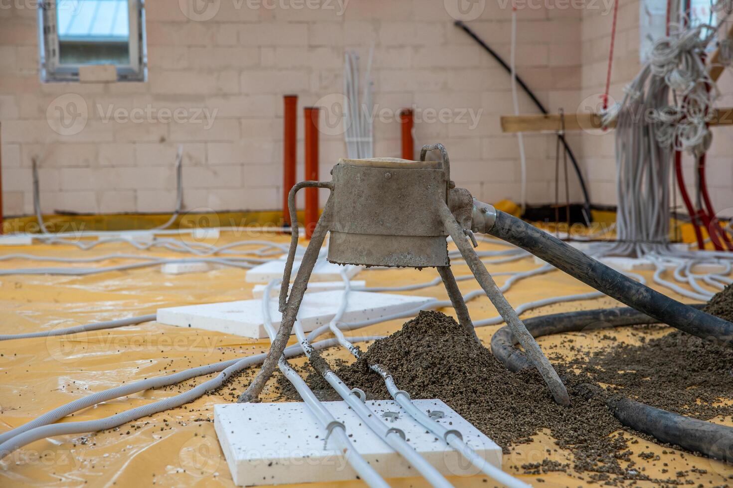 An indoor construction site featuring a concrete screed pump, yellow protective sheet, pipes, conduits, light brick walls, and scattered tools. photo