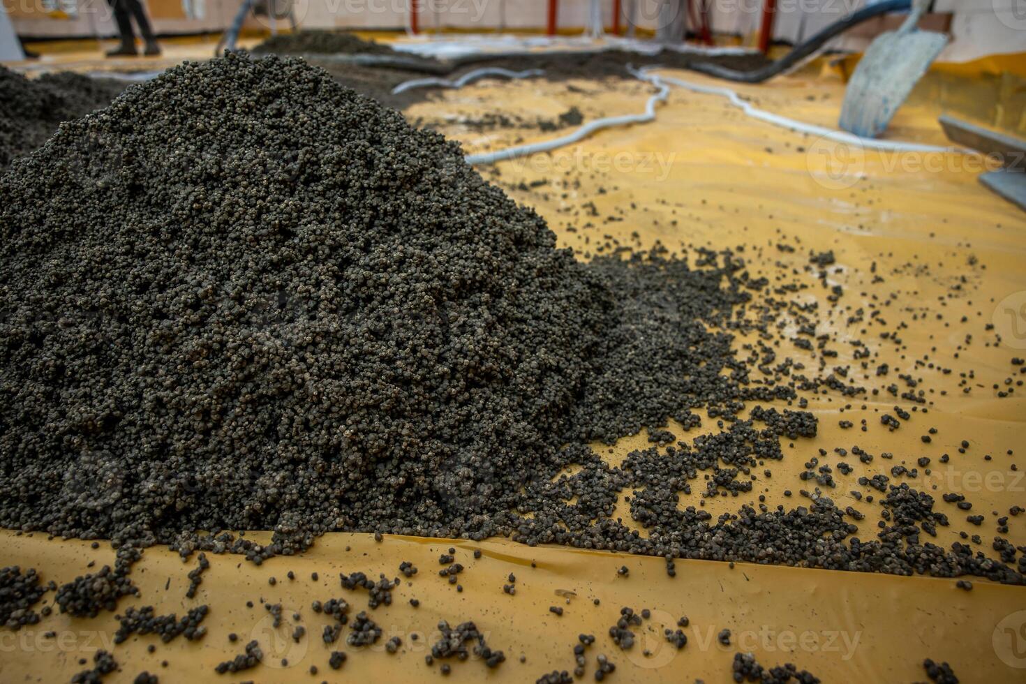 Dark granular material spread on a yellow surface in an indoor industrial facility, with visible pipes, hoses, and equipment in the background. photo