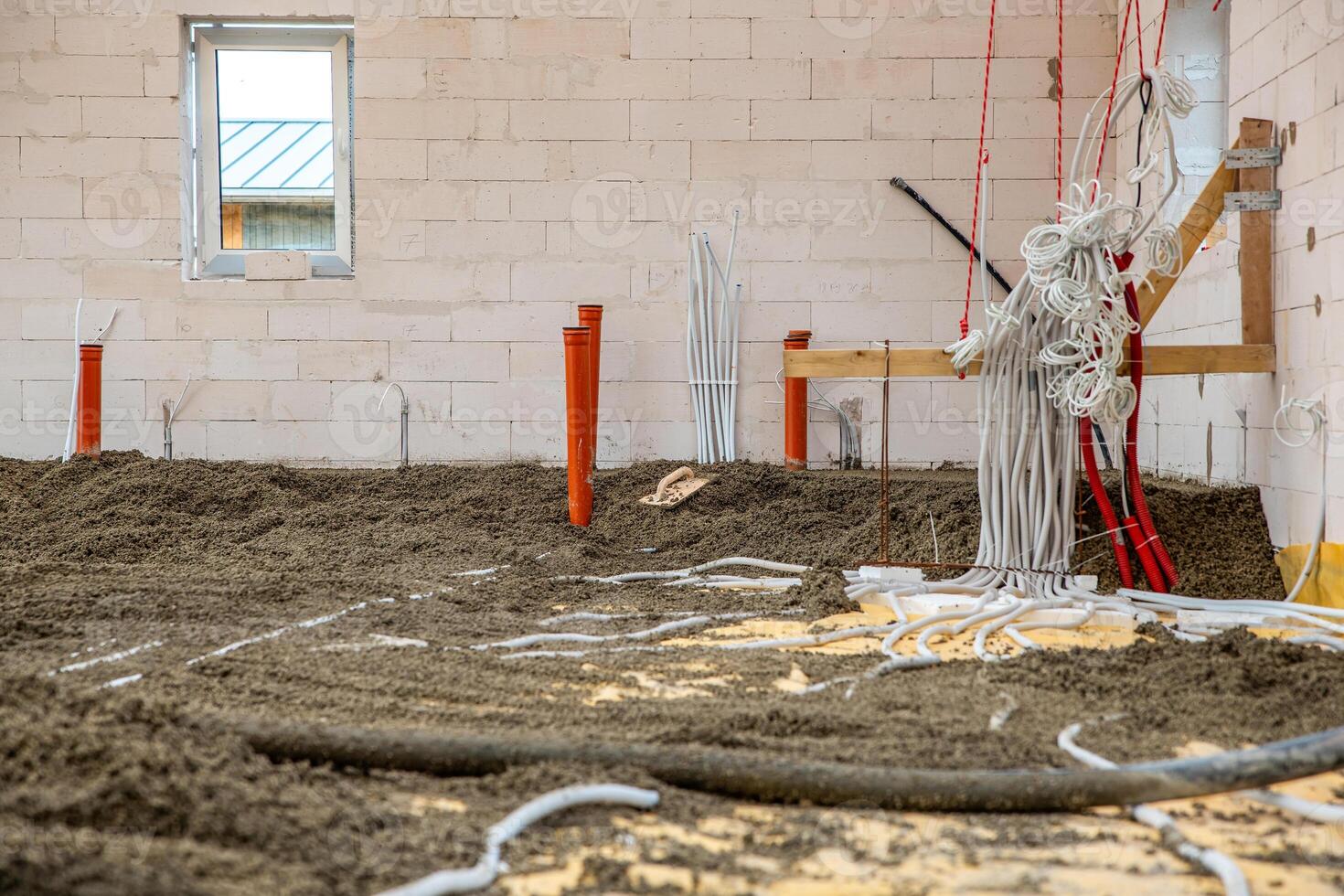 Construction site interior showing light brick walls, a window with a metal roof outside, exposed pipes, loose material on the floor, and wooden supports. photo