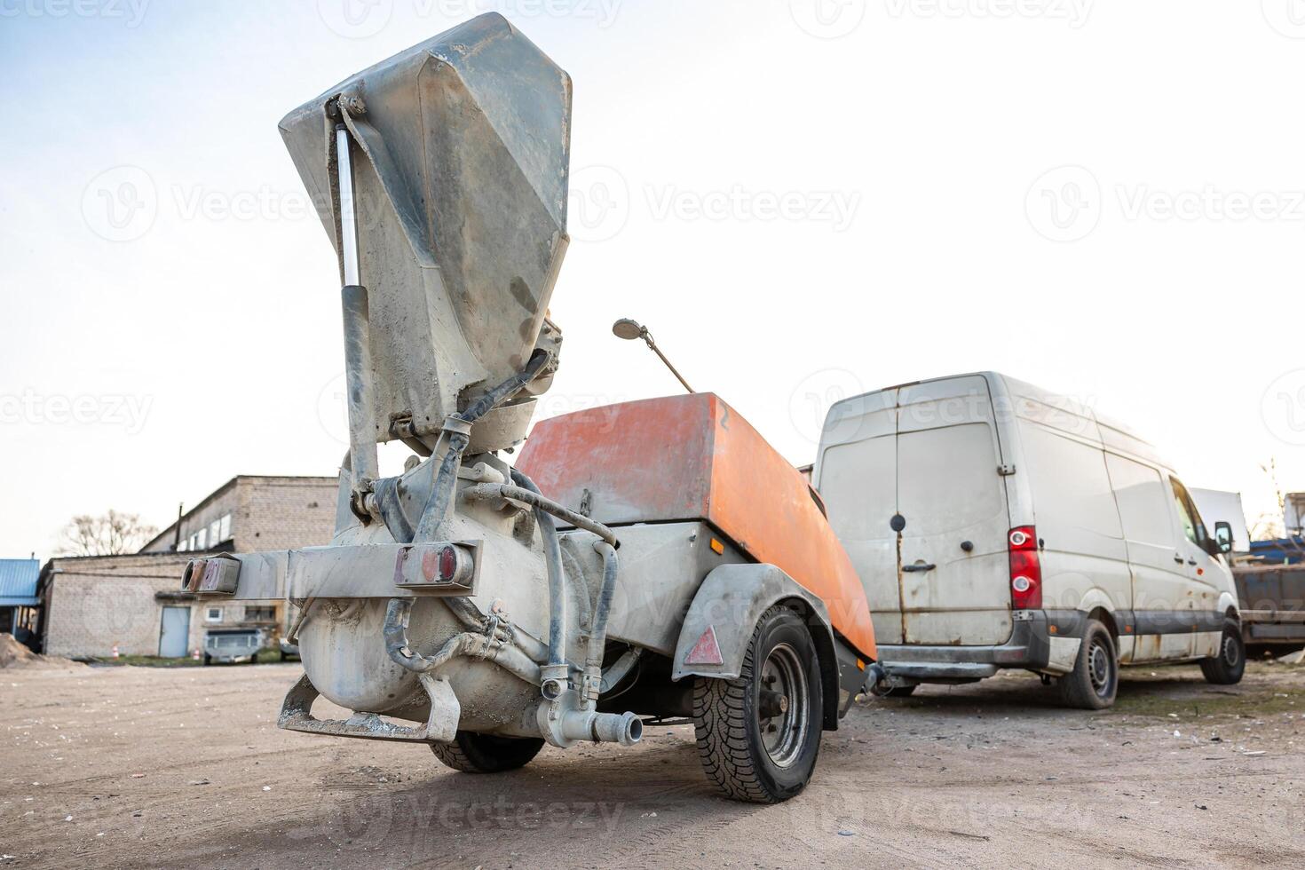 A concrete pump trailer with an orange and gray body stands near a white van on dirt ground, surrounded by industrial buildings and vehicles. photo