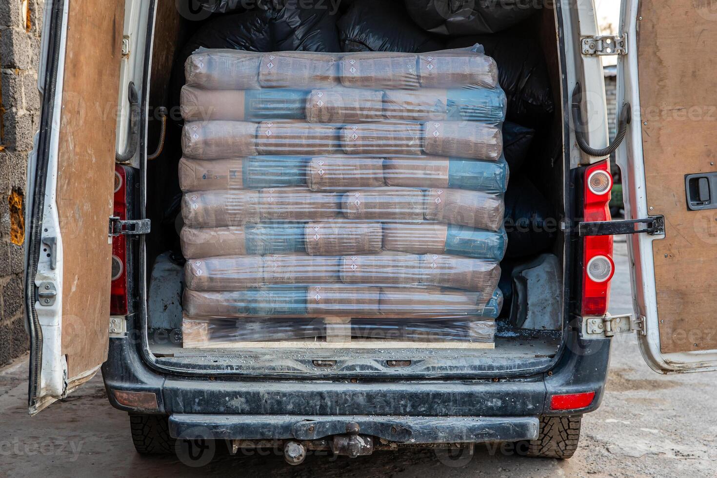 A stack of plastic wrapped cement bags on a wooden pallet inside a white delivery van. Black bags fill the rear space. Van parked on concrete near cinder block wall. photo