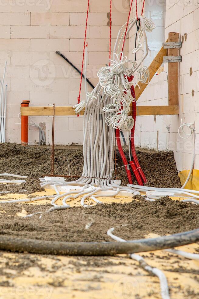 An indoor construction site featuring white brick walls, coiled electrical conduits, red ropes, wooden braces, and a floor with loose material and pipes. photo