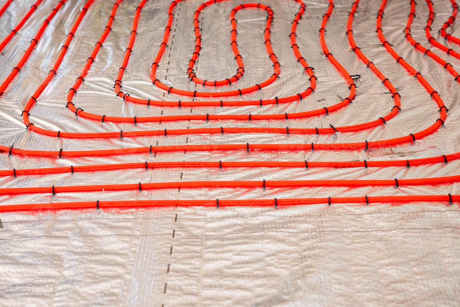 Bright red underfloor heating pipes arranged in loops, secured with black fasteners on a metallic insulation layer, ready for Estrich concrete application. photo