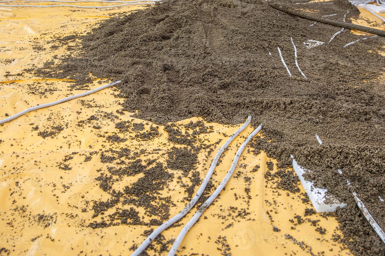 A construction site showing a yellow plastic sheet partially covered by gravel or concrete mix, with white pipes scattered across the surface. photo