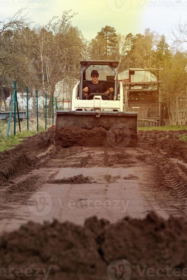 A man uses a small bulldozer to level a dirt path. Trees and fencing border the area, with freshly turned soil in the foreground and a structure visible. photo