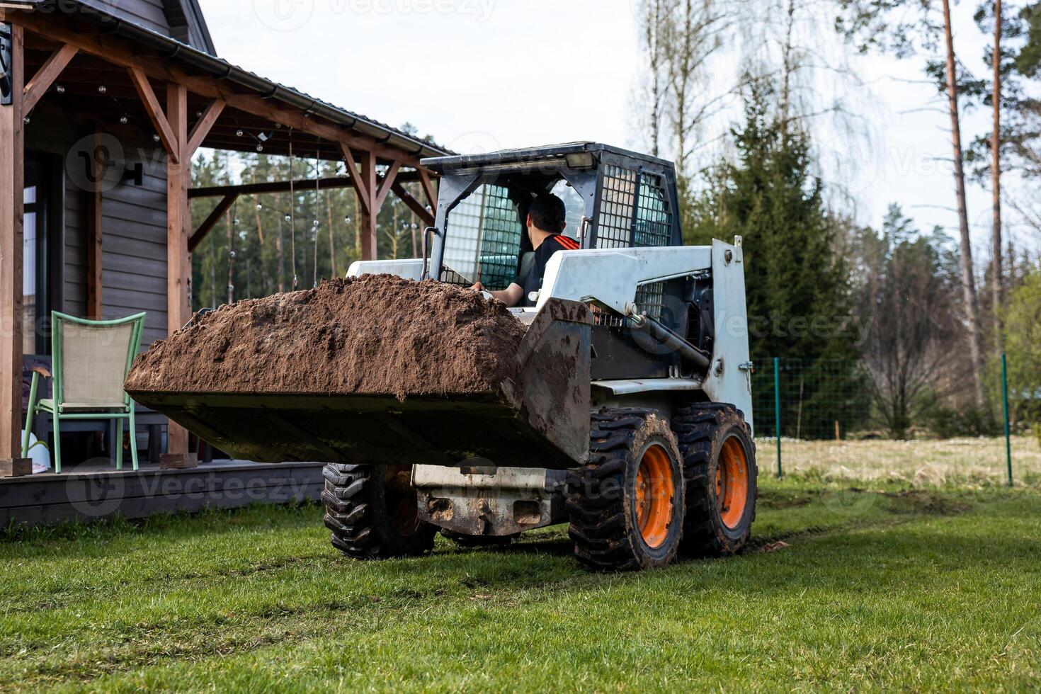 A skid steer loader with orange wheels moves soil on grass near a wooden house with a covered porch, chairs, string lights, trees, and a fenced area. photo