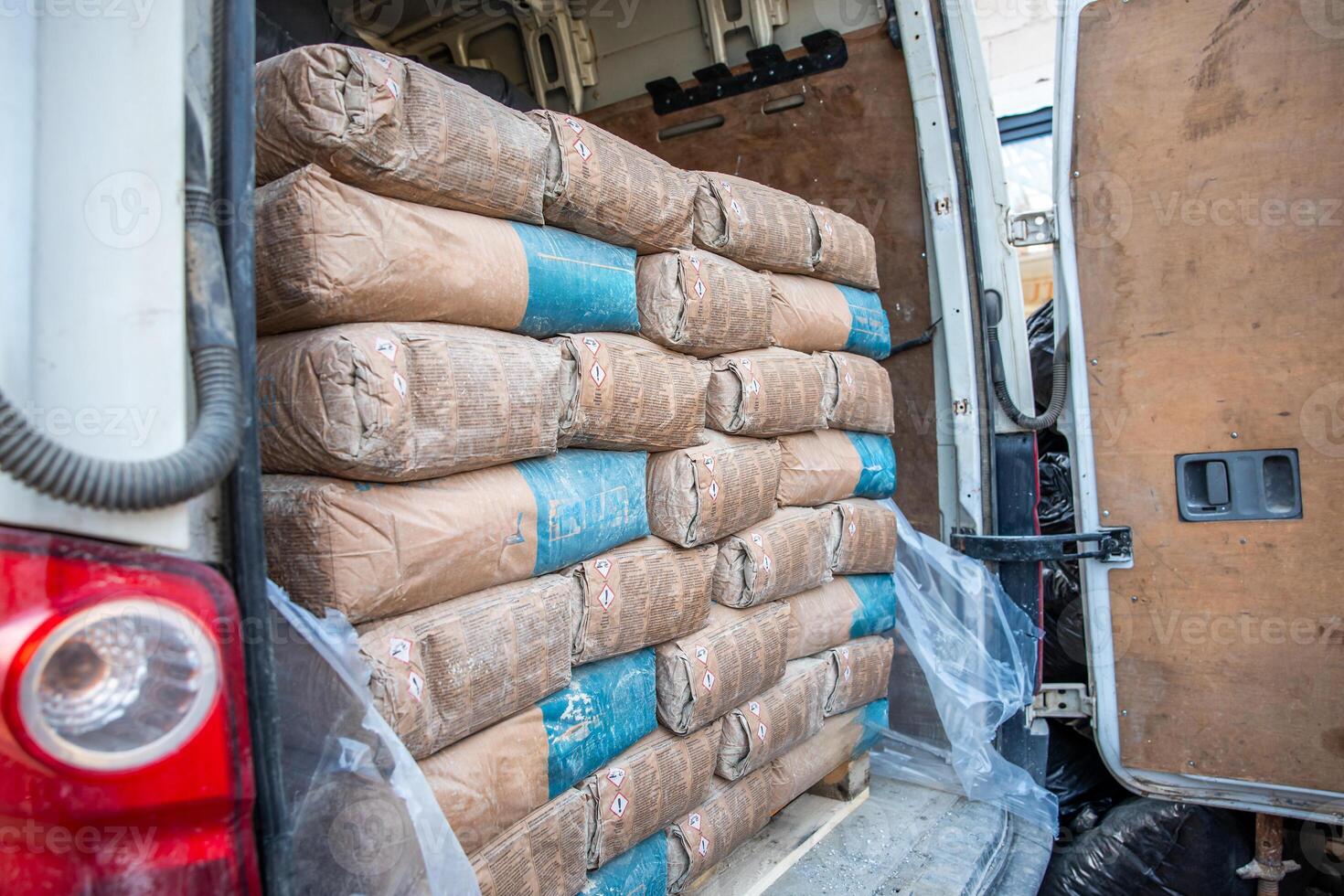 Open rear compartment of a white van with brown and blue cement bags stacked neatly. Wooden paneling and red taillight are visible, with plastic sheeting partially covering the cargo. photo