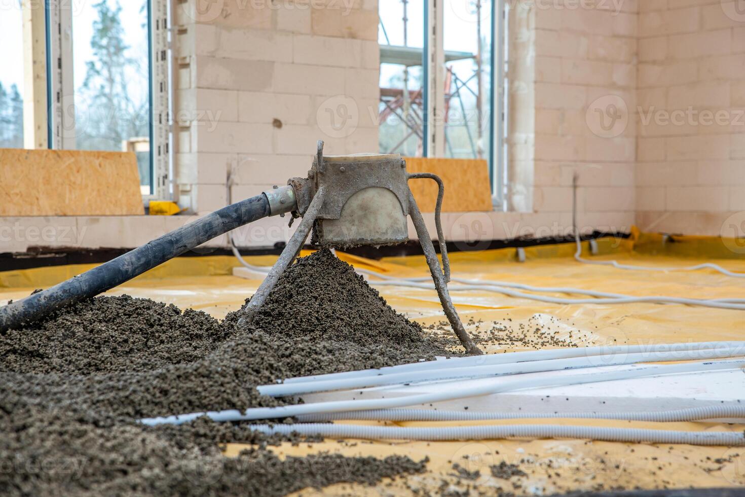 A construction site showing a concrete pump discharging onto a yellow sheet, white conduit pipes in the foreground, and scaffolding around white block walls. photo